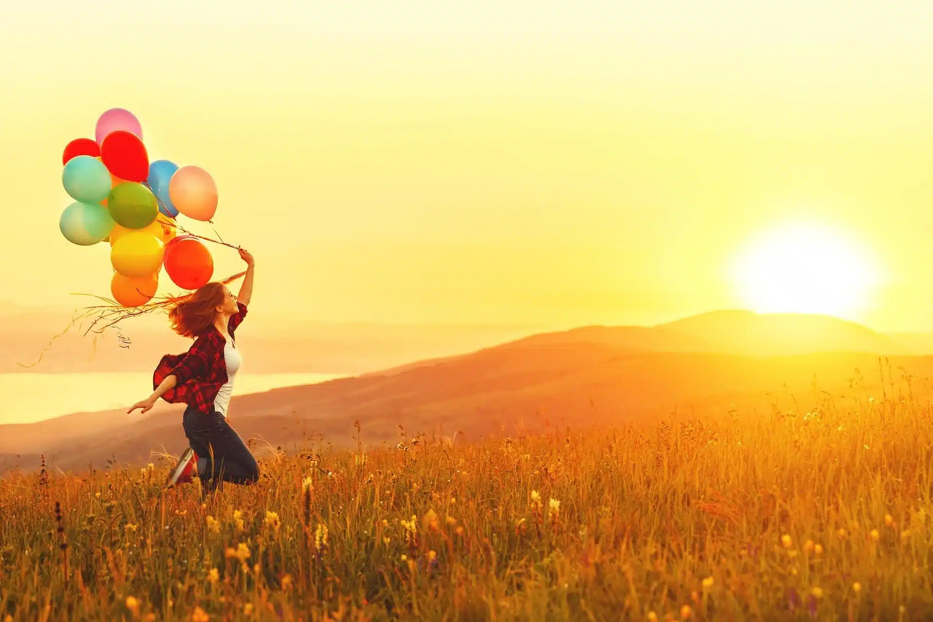 Happy cheerful girl with balloons running across meadow at sunset